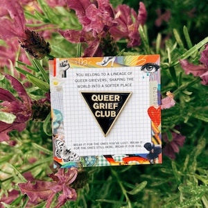 May include: A pin with the text "QUEER GRIEF CLUB" on a black triangle, set against a backdrop of lavender and greenery. The pin is on a card with the text "YOU BELONG TO A LINEAGE OF QUEER GRIEVERS, SHAPING THE WORLD INTO A SOFTER PLACE."