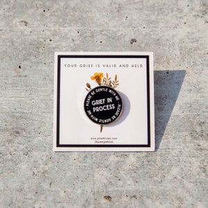 May include: A circular black and white enamel pin with the words "GRIEF IN PROCESS". The pin is on a white card with the text "YOUR GRIEF IS VALID AND HELD". The card has a black border and a small floral arrangement.