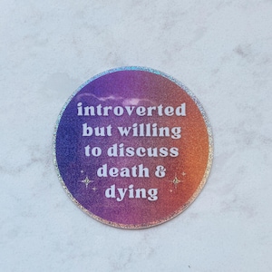 May include: A round sticker with a gradient of purple, orange, and gold. The text "introverted but willing to discuss death & dying" is in white. The sticker has a glittery silver border.