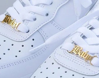 May include: A pair of white leather sneakers with white laces and gold-colored "Baby" lace charms. The shoes have a classic design with perforations on the toe box and a white sole. The background is blurred, suggesting an outdoor setting.