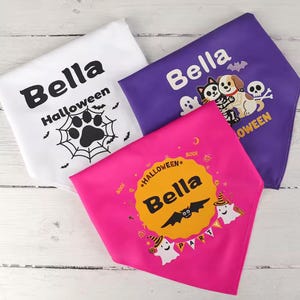 May include: Three Halloween-themed dog bandanas in white, purple, and pink. The white bandana has a paw print and spiderweb design. The purple bandana features a cartoon dog, cat, and skull. The pink bandana has a bat and ghost design. All bandanas have the name "Bella".