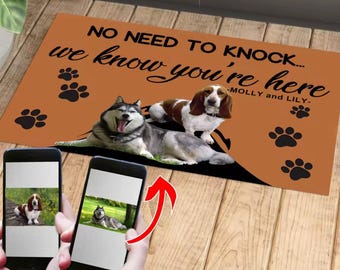 以下が含まれることがあります： 「NO NEED TO KNOCK... we know you're here」の文字と足跡がプリントされたオレンジ色のドアマット。シベリアンハスキーとバセットハウンドが描かれています。「Molly and Lily」の名前でパーソナライズされています。