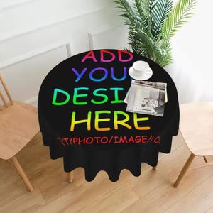 May include: A round black tablecloth with the text "ADD YOUR DESIGN HERE" in rainbow colors. A white teacup and a magazine are on the table. The tablecloth is on a table with wooden legs.