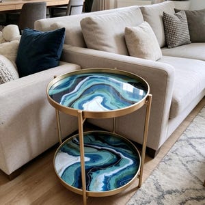 May include: A two-tier round side table with a gold-toned metal frame. Both the top and bottom circular shelves feature a glossy, resin-like surface with a swirling, marbled pattern in shades of deep navy blue, teal, turquoise, and white.