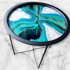 May include: A circular side table with a black metal frame and a decorative top. The tabletop has an abstract design in teal, white, and gold, reminiscent of a geode. The table stands on a three-legged base.