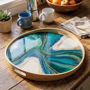 May include: Circular wooden serving tray with a resin top. The resin design features teal, white, and gold, resembling an abstract agate pattern. The tray has built-in handles and is displayed on a wooden surface.