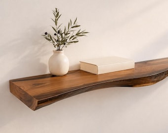May include: A live edge wooden floating shelf with a rich brown grain pattern. On the shelf sits a small, cream-colored ceramic vase containing a green olive branch with dark olives, and a single rectangular book with a plain, light-colored cover.