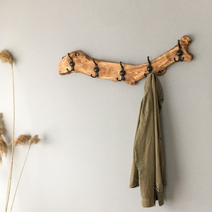 May include: A rustic wooden coat rack with six black metal hooks. The rack is made from a natural, light brown branch and is mounted on a gray wall. An olive green jacket hangs from one of the hooks. Decorative dried plants are in a wooden vase.