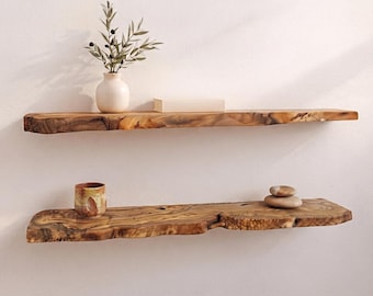 Op de afbeelding: Twee horizontale wandplanken van natuurlijk hout met boomkanten. De bovenste plank bevat een witte keramische vaas met een groene olijftak en een rechthoekig boek. De onderste plank bevat een kleine keramische beker en twee gestapelde stenen.