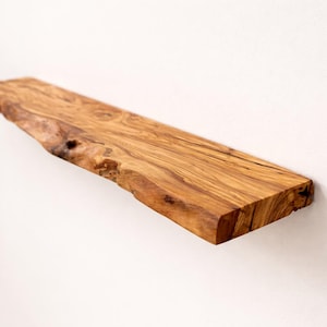May include: A rectangular wall shelf crafted from natural olive wood, featuring a prominent live edge with a raw, textured bark finish on the front side. The wood displays a rich, golden-brown grain pattern with dark, swirling knots.