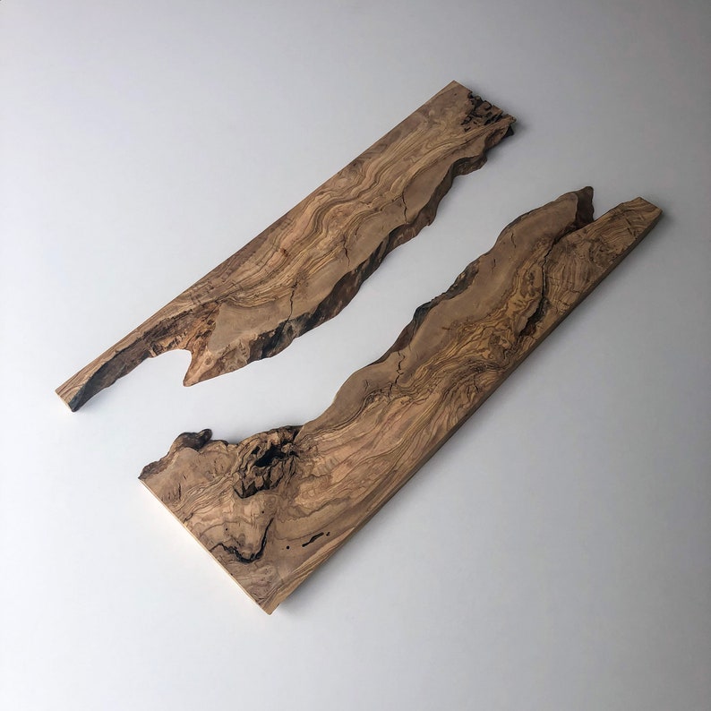 May include: Two olive wood slabs with natural, live edges. The wood grain features a mix of light and dark brown tones. The slabs are rectangular, with one having a unique cutout. Ideal for woodworking projects or decorative use.