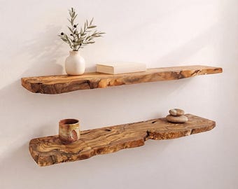 May include: Two horizontal wall-mounted shelves made from natural live-edge wood with visible grain patterns. The top shelf holds a white ceramic vase with a green plant and a closed book. The bottom shelf holds a small ceramic cup and two stacked stones.