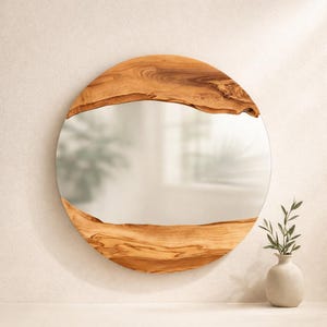 May include: Round mirror with a natural wood frame. The frame is made of light brown wood with a unique grain pattern. The mirror is circular and reflects the surrounding environment. A small, white vase with greenery is in the foreground.