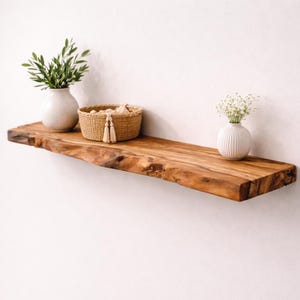 May include: A live edge wooden floating shelf holding a light-colored ceramic vase with green foliage, a round woven basket with two hanging tassels, and a small white ribbed ceramic vase containing delicate white flowers against a plain wall.
