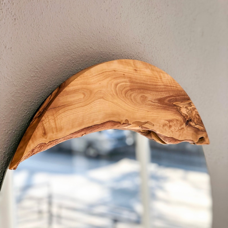 May include: A wooden mirror with a natural, organic design. The frame is crafted from a single piece of light brown wood, showcasing the grain and texture. The mirror has a curved, arched shape, adding a rustic and elegant touch to any space.