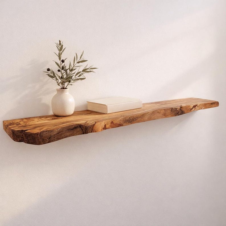 May include: A live edge wooden floating shelf with a natural grain pattern and rough bark edges. A small, cream-colored ceramic vase containing a green olive branch with dark olives and a rectangular, cream-colored book sit on the shelf surface.