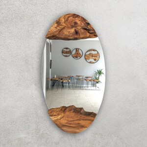 May include: Oval-shaped mirror with a natural wood frame. The frame is made of a light brown, textured wood, with the grain visible. The mirror reflects a room with wooden tables and stools.