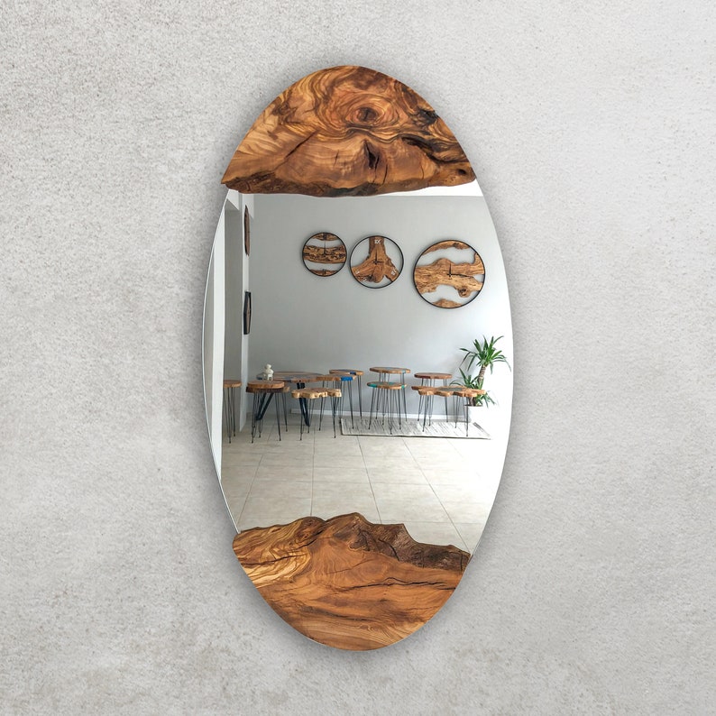 May include: Oval-shaped mirror with a natural wood frame. The frame is made of a light brown, textured wood, with the grain visible. The mirror reflects a room with wooden tables and stools.
