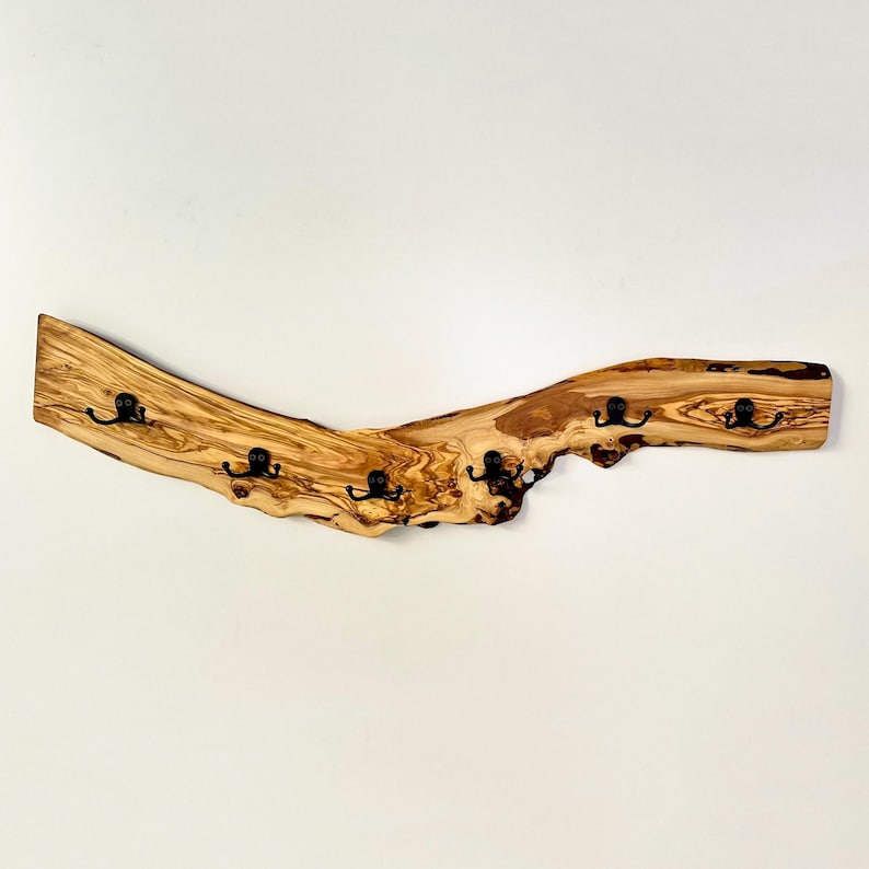 May include: A wooden coat rack with five black metal hooks. The rack is made from a piece of light brown wood with a natural, curved shape. The wood has a visible grain and knots, adding to its rustic appeal. The hooks are evenly spaced along the bottom edge.