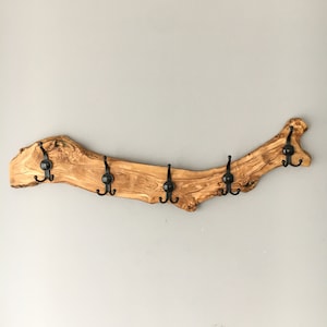 May include: A rustic wooden coat rack made from a natural olive branch. It features six black metal hooks evenly spaced along the curved, light brown wood. The wood grain is visible, adding to the natural aesthetic. A decorative and functional wall hanging.