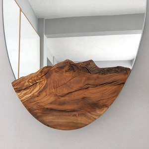 May include: A round mirror with a natural wood accent. The wood is a warm brown color and has a mountain-like shape. The mirror is mounted on a light gray wall.