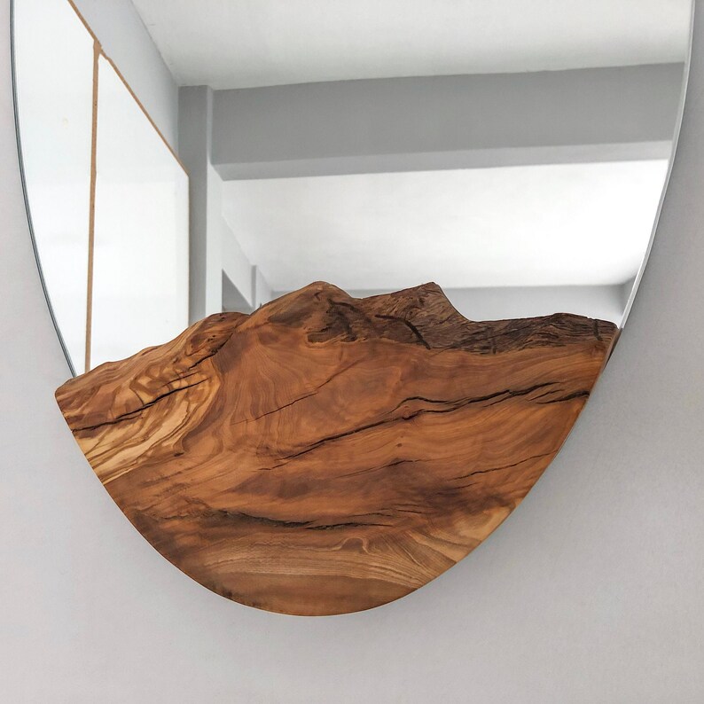 May include: A round mirror with a natural wood accent. The wood is a warm brown color and has a mountain-like shape. The mirror is mounted on a light gray wall.