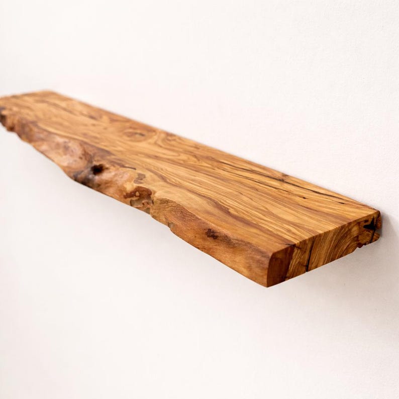 May include: A wall-mounted floating shelf crafted from natural olive wood, featuring a prominent live edge with an irregular, organic contour. The wood displays a rich, golden-brown grain pattern with dark, swirling knots and a smooth finish.