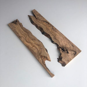 May include: Two irregularly shaped wooden planks with natural edges. The planks are a warm brown color with darker grain patterns. The wood appears to be olive wood, with a smooth, polished finish. The planks are set against a white background.