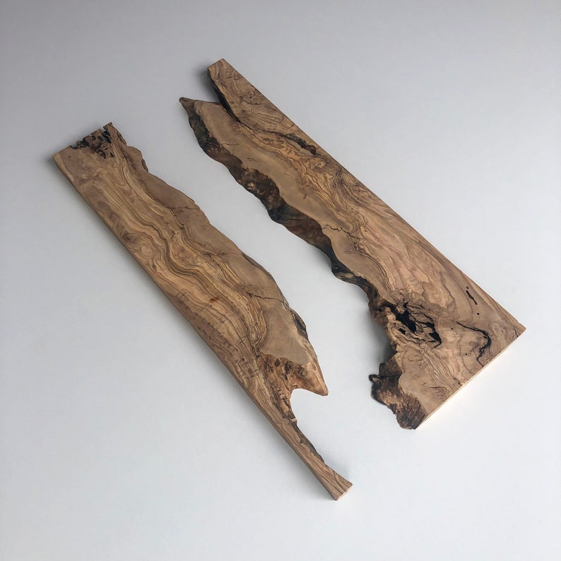 May include: Two irregularly shaped wooden planks with natural edges. The planks are a warm brown color with darker grain patterns. The wood appears to be olive wood, with a smooth, polished finish. The planks are set against a white background.