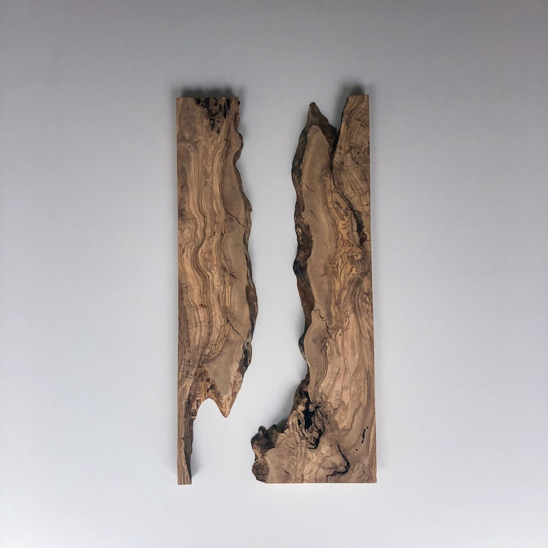 May include: Two vertical wooden panels with irregular edges, showcasing a natural wood grain pattern in shades of brown and tan. The panels are mounted against a plain, light gray wall, creating a minimalist aesthetic.