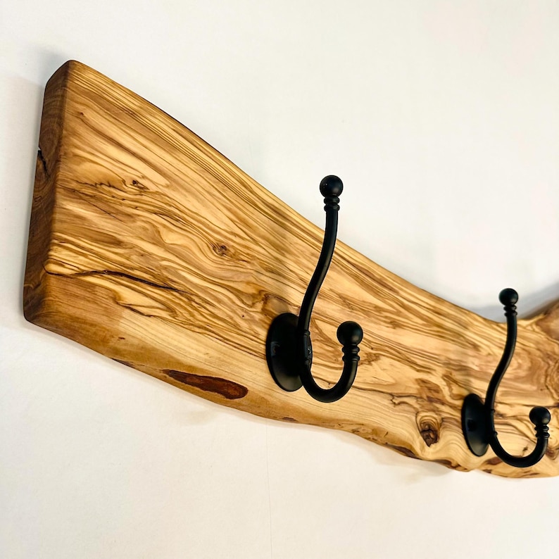 May include: A wooden coat rack with three black metal hooks. The rack is made from a light-colored, textured piece of wood with a natural edge. The hooks are curved and have a rounded ball at the end.