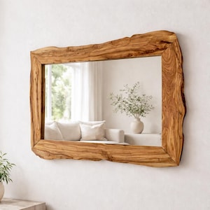 Op de afbeelding: Een rechthoekige spiegel met een natuurlijke houten lijst. De lijst heeft een ruwe, organische rand, die de nerf en textuur van het hout laat zien. De spiegel reflecteert een lichte kamer met een witte bank en een vaas met bloemen.