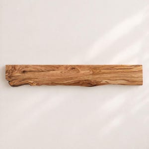 May include: A long, rectangular wooden serving board crafted from olive wood, featuring a natural live edge on the left side. The surface displays a prominent, swirling grain pattern in shades of light and dark brown, highlighting the wood's texture.