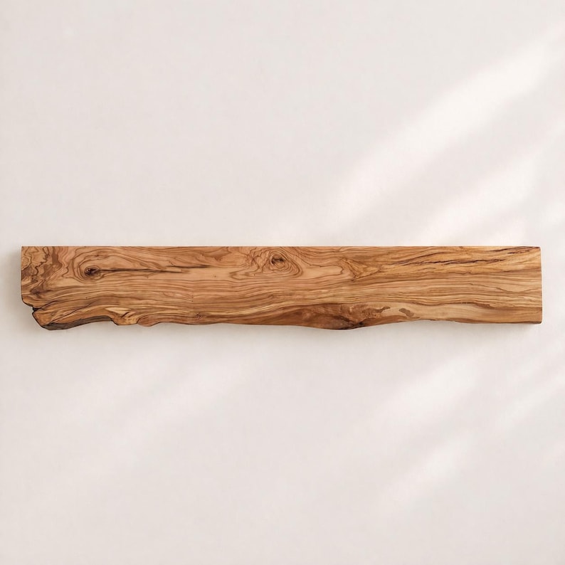 May include: A long, rectangular wooden serving board crafted from olive wood, featuring a natural live edge on the left side. The surface displays a prominent, swirling grain pattern in shades of light and dark brown, highlighting the wood's texture.