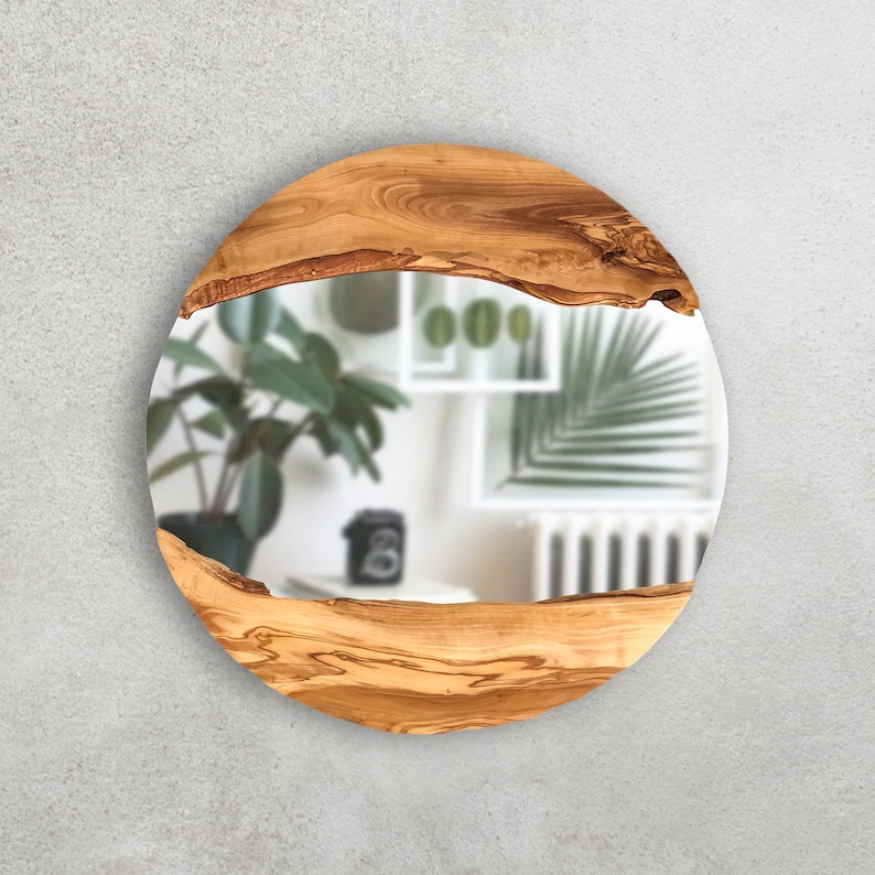 May include: Round mirror with a natural wood frame. The frame is made of light brown wood with a unique grain pattern. The mirror reflects a blurred interior scene with plants and a white shelf.