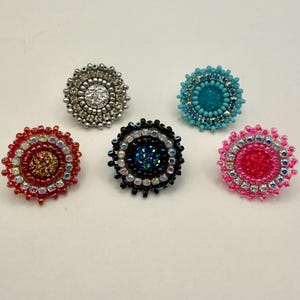 Könnte beinhalten: Eine Sammlung von fünf Perlenringen in verschiedenen Farben. Die Ringe haben ein zentrales Design, das von einem Perlenrand umgeben ist. Farben sind Silber, Türkis, Rot, Schwarz und Pink. Die Ringe sind wahrscheinlich handgefertigt.
