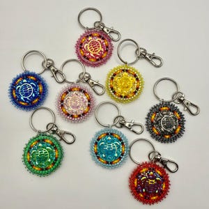 May include: A selection of beaded keyrings in shades of pink, blue, green, yellow, and red. Each keyring showcases a turtle design at the centre, encircled by detailed beadwork and a silver-coloured clasp.