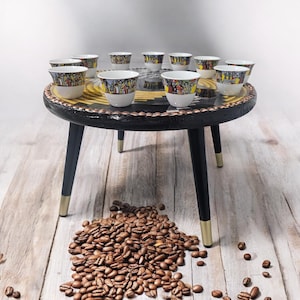 May include: A black, round side table with a decorative top and black legs with gold tips. The table holds several small white coffee cups with colorful designs. A pile of coffee beans rests on the wooden floor.