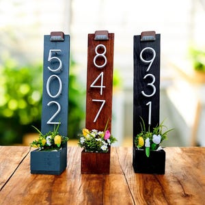 May include: Three wooden address signs in blue, brown, and black, each with white numbers and a small planter box filled with flowers. The signs have a rustic, farmhouse style and are displayed on a wooden surface.