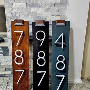 May include: Three wooden house number signs with solar powered lights. The numbers are 787, 987, and 487. The signs are painted in brown, black, and blue.