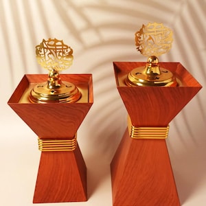 May include: Two wooden incense burners with gold accents. Each has a square top with a gold interior, a gold base, and a gold decorative top with Arabic script. The wood has a reddish-brown finish.