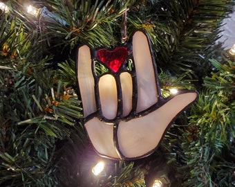 May include: A stained glass ornament shaped like a hand signing "I love you" in American Sign Language. The hand is white with black outlines and a red heart in the center.