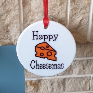 May include: A white ceramic bauble with a red ribbon hanging from it. The bauble has the text "Happy Cheesemas" and a cartoon image of a wedge of cheese.
