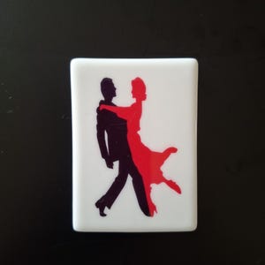May include: A rectangular white tile featuring a graphic silhouette of two figures dancing. One figure is rendered in solid black, while the other figure is rendered in solid red. The two figures are positioned in a close embrace, mid-dance.