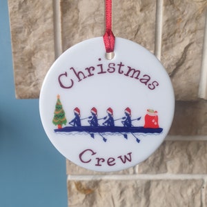 May include: White ceramic Christmas bauble with a red ribbon, depicting a group of rowers in a boat with Santa's sack and a Christmas tree. The text "Christmas Crew" is printed on the bauble.