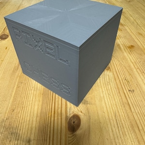 Peut inclure: Un cube gris avec le mot "PIXEL" imprimé sur le côté. Le cube est fait d'un matériau texturé et a une surface légèrement rugueuse.