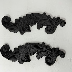 May include: Two identical black decorative appliques featuring an intricate, elongated acanthus leaf scroll design. Each piece has a curved, symmetrical shape with detailed, raised foliage patterns that spiral at both ends, finished in a matte black.