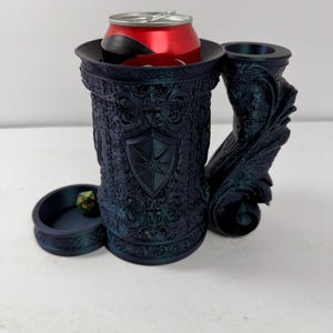 May include: A dark blue, textured can holder shaped like a tankard with an ornate shield emblem on the front and a large, sculpted handle. A red soda can is inserted into the top, and a small, separate circular tray holds a green multi-sided die.