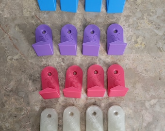 Puede incluir: Un conjunto de 12 soportes de pared en cuatro colores diferentes: azul claro, morado, rosa y blanco. Cada soporte tiene un diseño único con un pequeño círculo en el centro.