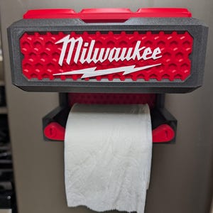 Może przedstawiać: Ścienny uchwyt na papier toaletowy z czarno-czerwoną ramą o strukturze plastra miodu. W górnej części znajduje się białe logo Milwaukee z grafiką błyskawicy. Na uchwycie zawieszona jest standardowa rolka białego papieru toaletowego.
